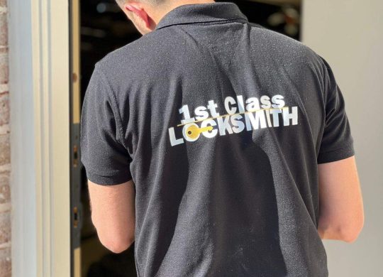 1st Class Locksmith technician working on a residential door lock, wearing a black uniform with yellow company branding in Prosper, TX